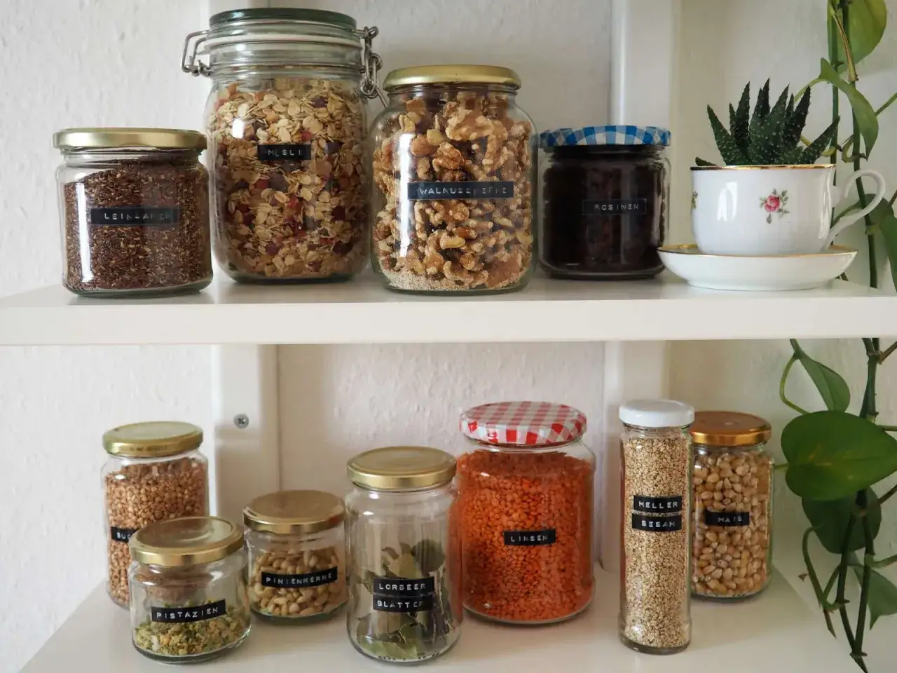 Regale mit Gläsern, die mit Etiketten versehen sind, um den Inhalt zu kennzeichnen: Müsli, Walnüsse, Leinsamen, Linsen, Mais, Sesam, Pistazien, Pinienkerne und Lorbeerblätter.