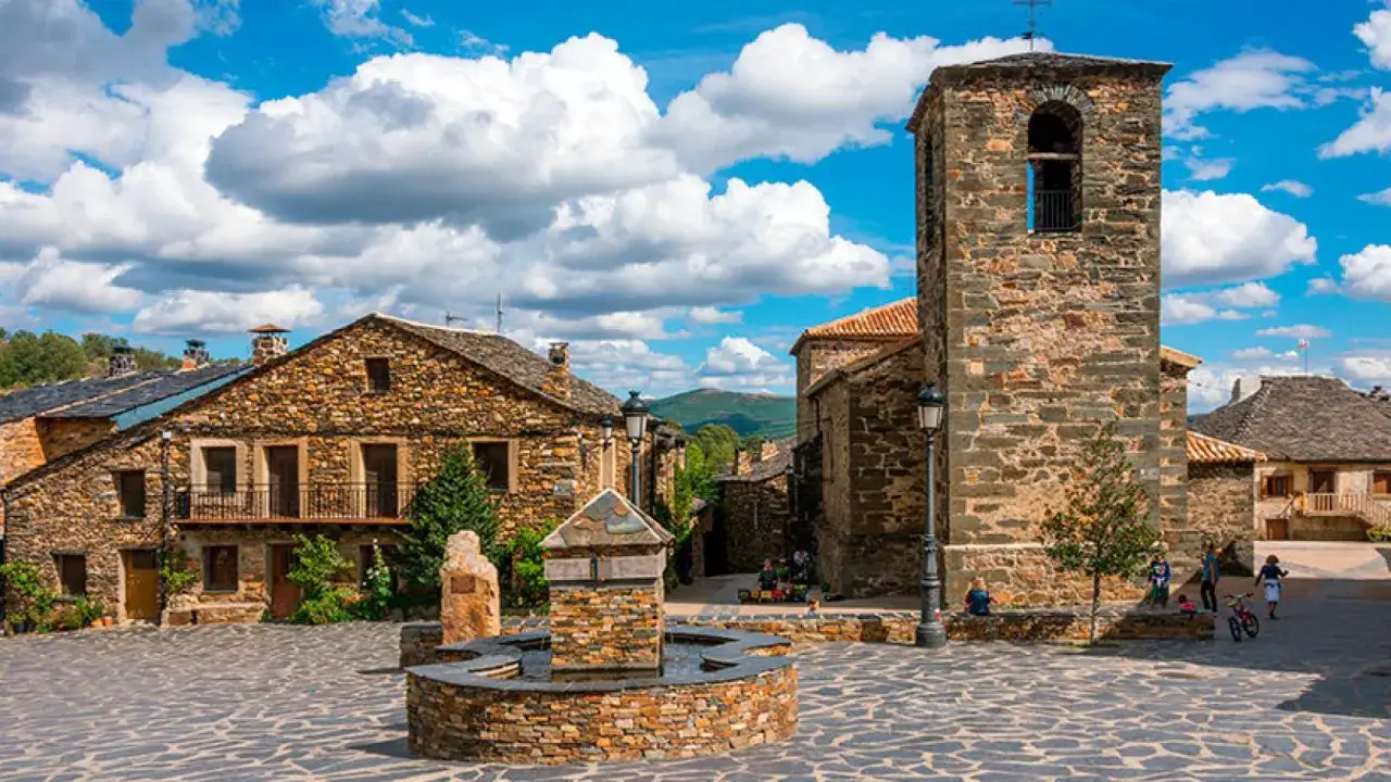 Plaza de pueblo con fuente y torre de iglesia de piedra. Un lugar encantador que ver en Castilla la Mancha, con casas de piedra y cielo azul.