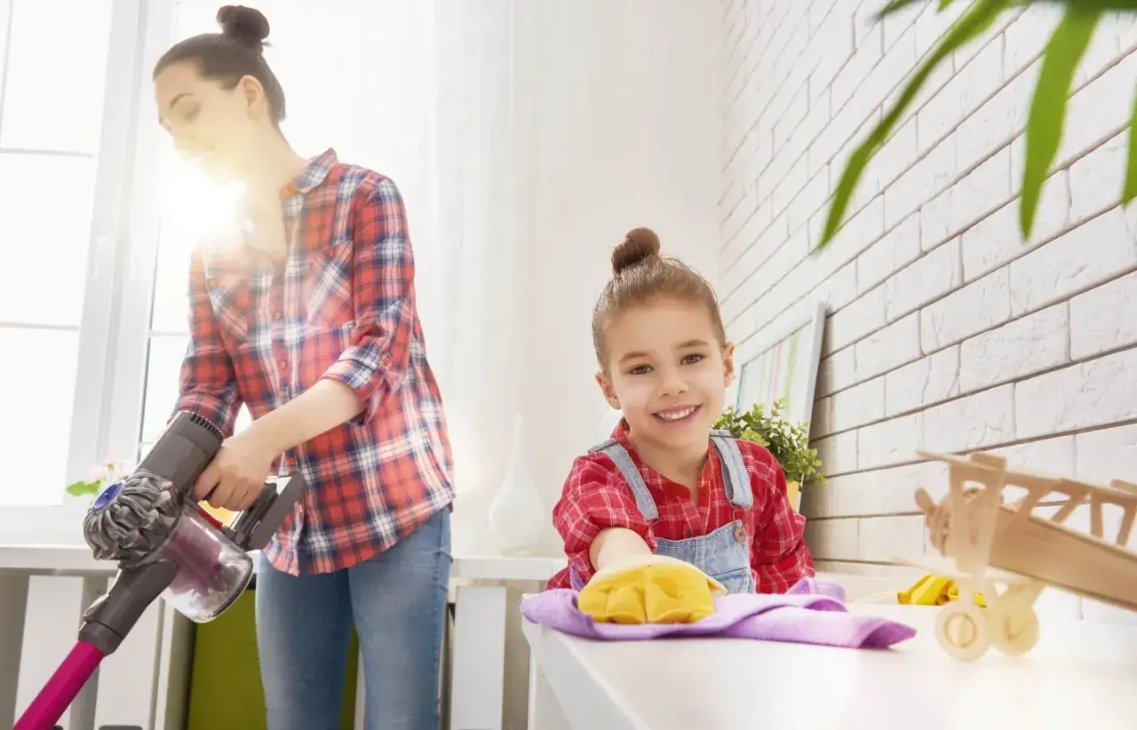 Mama odkurza, a 9-latka z uśmiechem pomaga w porządkach, wycierając kurze. Obowiązki 9 latka w domu to też nauka odpowiedzialności.