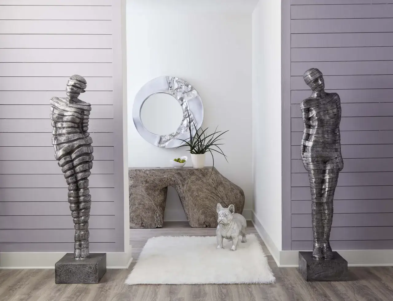 Dos esculturas de figuras humanas abstractas, de metal plateado, flanquean un pasillo con un espejo circular y un bulldog franc&eacute;s decorativo.