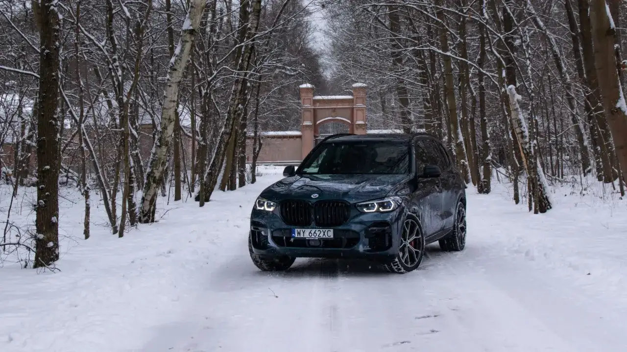 Ciemnozielone BMW X5 45e na śnieżnej drodze w zimowym lesie. W tle zabytkowa brama.