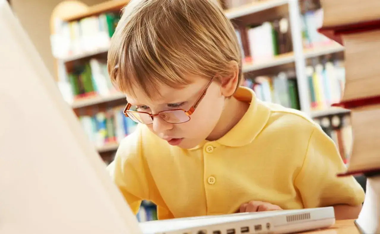 Chłopiec w okularach pochyla się nad laptopem, szukając informacji w bibliotece online dla studentów.
