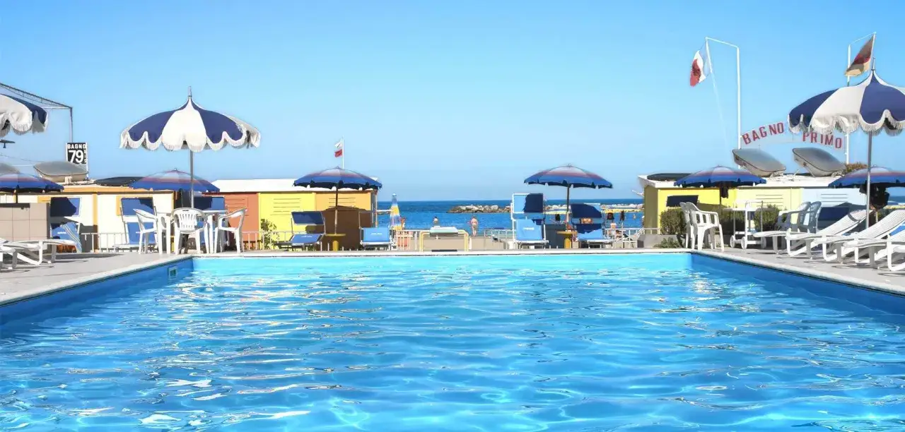 Piscina azzurra e lettini prendisole al **Valentini Family Village**, con cabine colorate e il mare sullo sfondo.