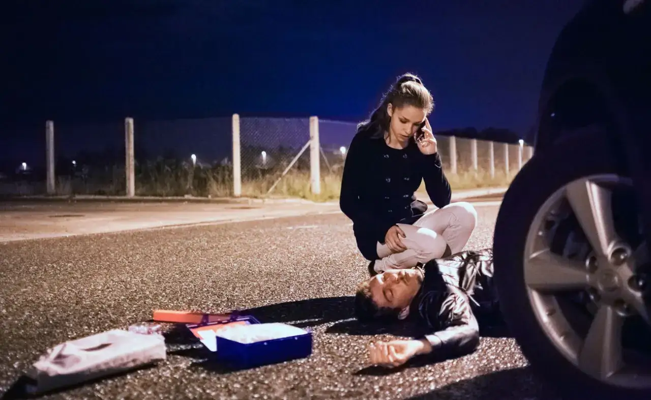 Una mujer llama por teléfono junto a un hombre tendido en el suelo, con un botiquín cerca. La primera medida a adoptar ante un accidentado es llamar a emergencias.