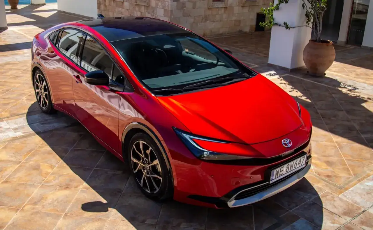 Nowy, czerwony Toyota Prius na tle śr&oacute;dziemnomorskiej architektury. Zastanawiasz się, ile pali hybryda? Ten model oferuje niskie spalanie.