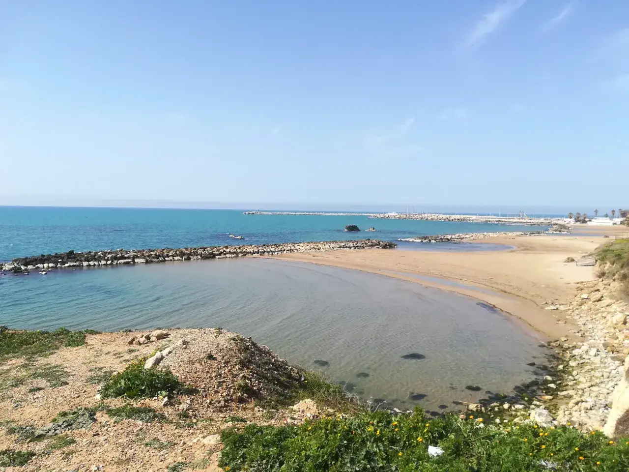Spiaggia dorata e mare cristallino, con dighe frangiflutti che creano piccole calette siciliane.