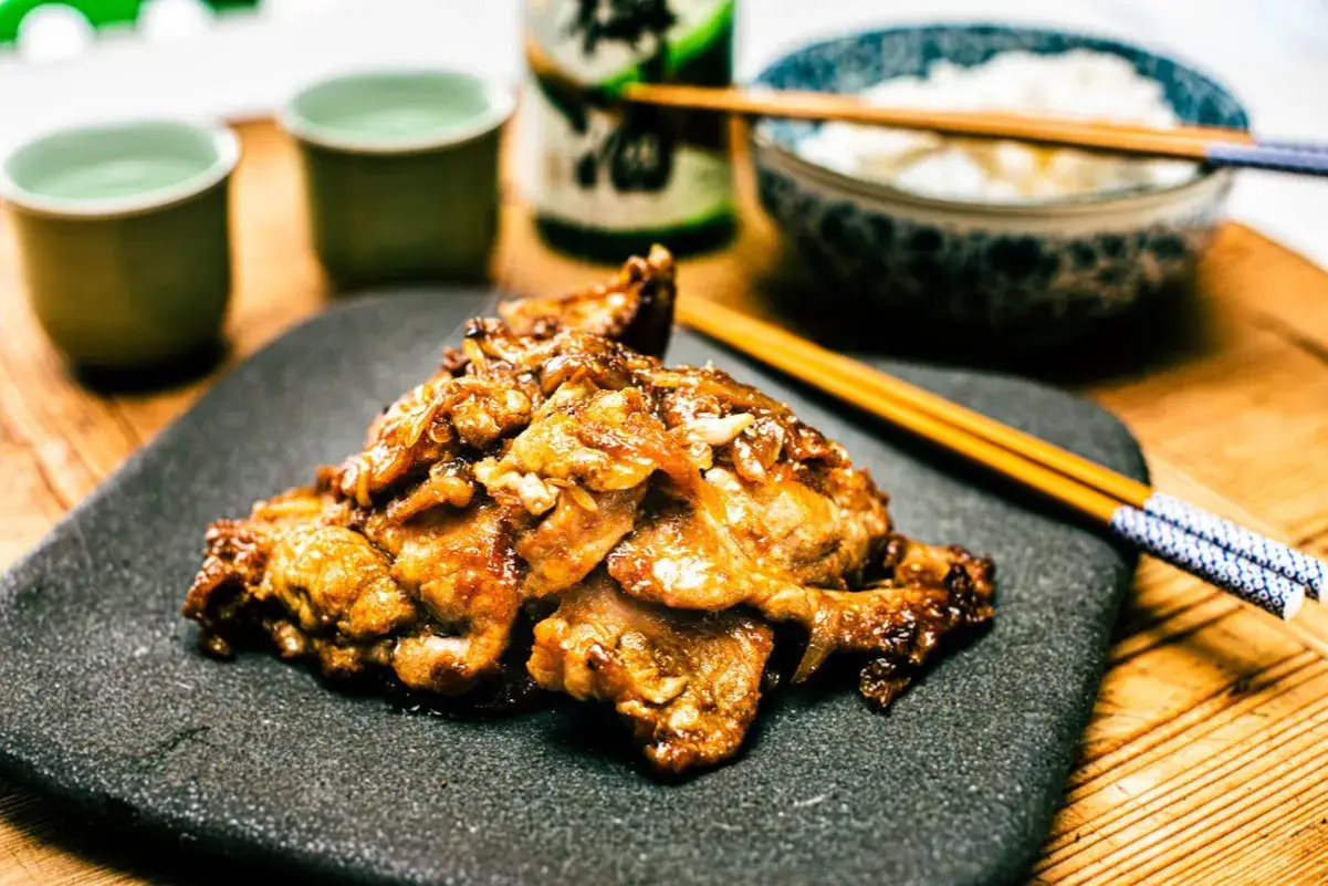 A plate of glistening pork shogayaki sits on a dark slate, ready to be enjoyed with rice and sake.