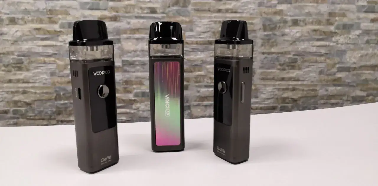 Tres dispositivos voopoo vinci pod, uno con un dise&ntilde;o iridiscente, sobre una superficie blanca y fondo de ladrillo.