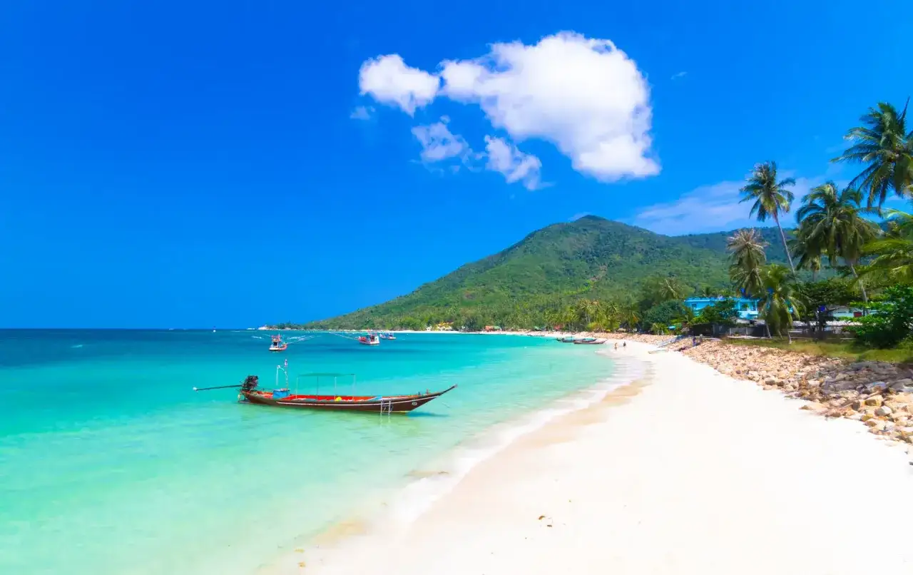 En vacker strand med turkost vatten, palmer och en traditionell båt vid Koh Phangan, Thailand.