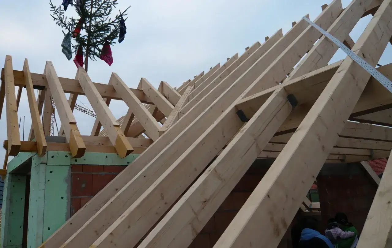 Der **Dachstuhlaufbau** schreitet voran. Holzbalken bilden das Gerüst, während oben ein geschmückter Baum mit bunten Tüchern steht.