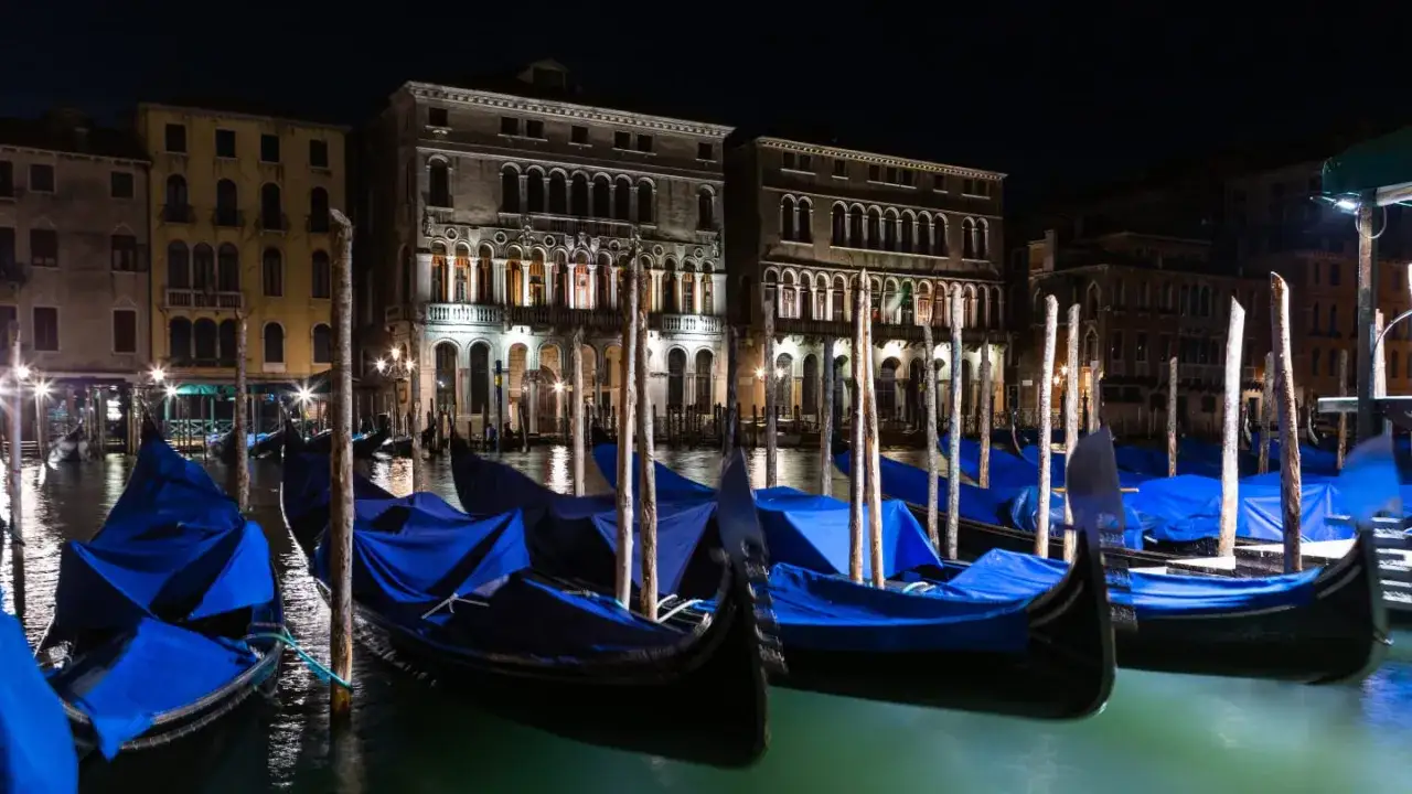Gondole a Venezia di notte, coperte da teli blu, ormeggiate davanti a palazzi illuminati.