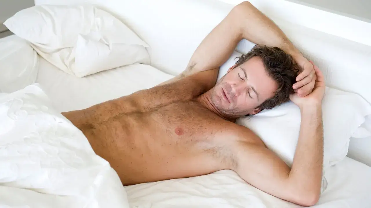 Hombre durmiendo plácidamente en una cama blanca, con los brazos cruzados sobre la cabeza. Se ve cómodo, quizás porque es bueno dormir sin ropa interior.