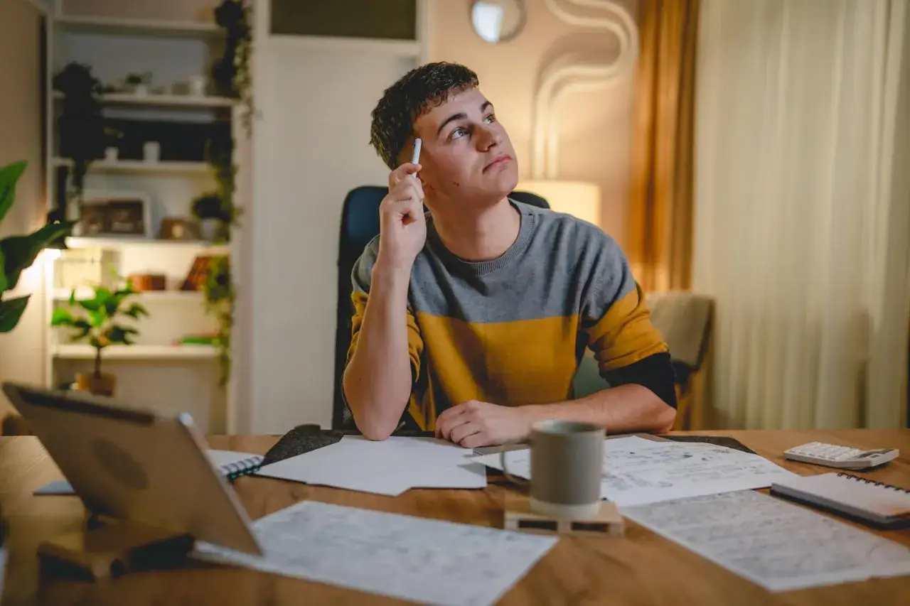 Un étudiant réfléchit, stylo à la main, devant sa table de travail. Il prépare comment commencer une soutenance de mémoire.