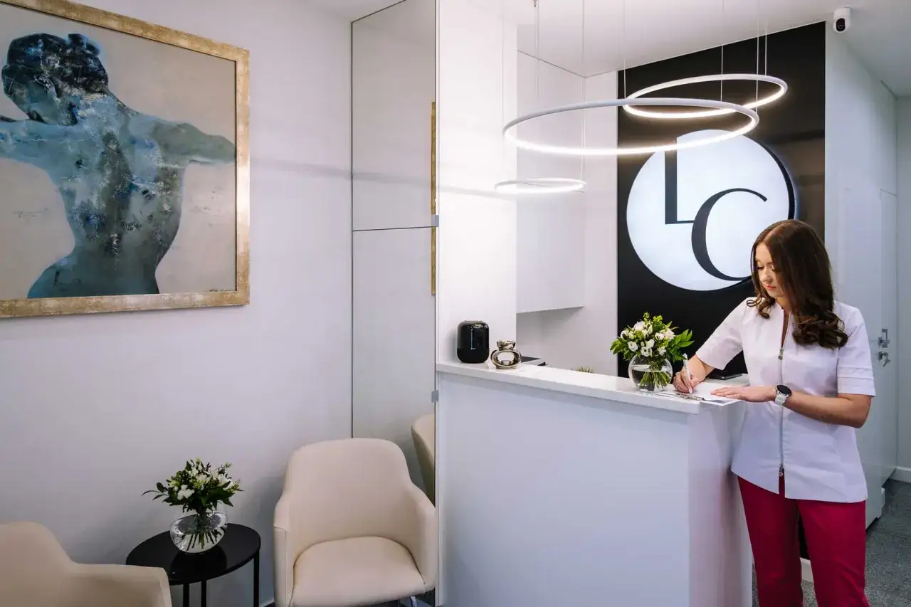 Recepcjonistka w klinice dermatologii estetycznej pisze coś na dokumentach przy białym kontuarze.