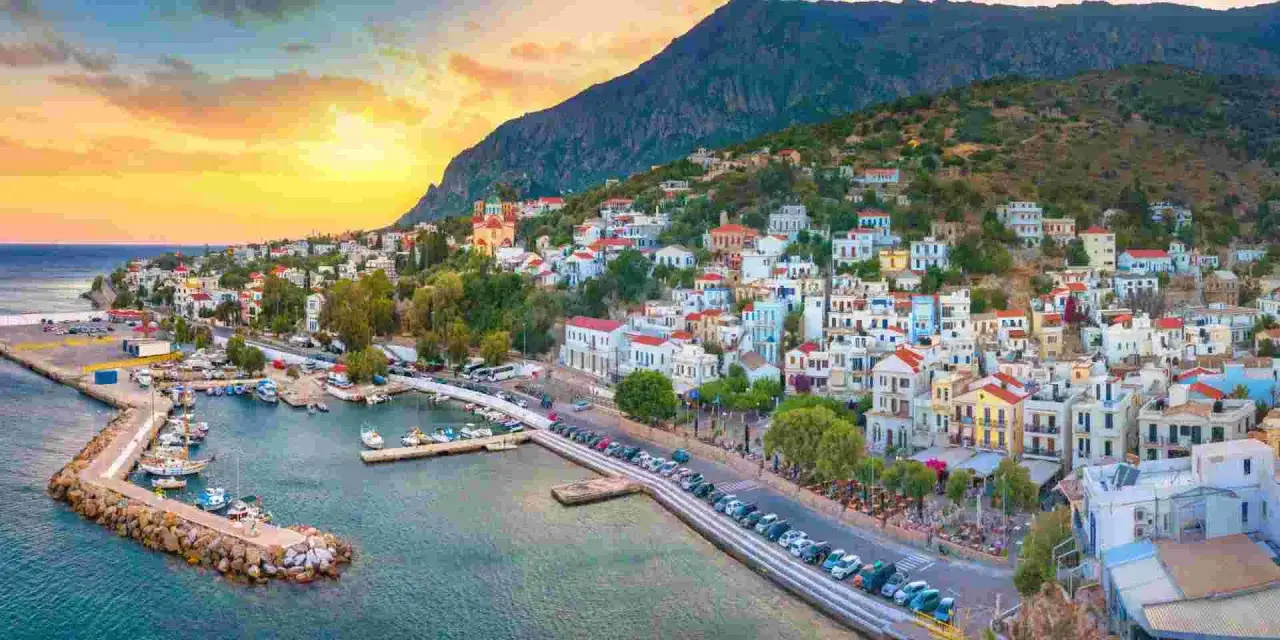 En pittoresk hamnstad på Ikaria, Grekland, med vita hus med röda tak som klättrar uppför en grönskande sluttning. Solnedgången färgar himlen i varma toner.