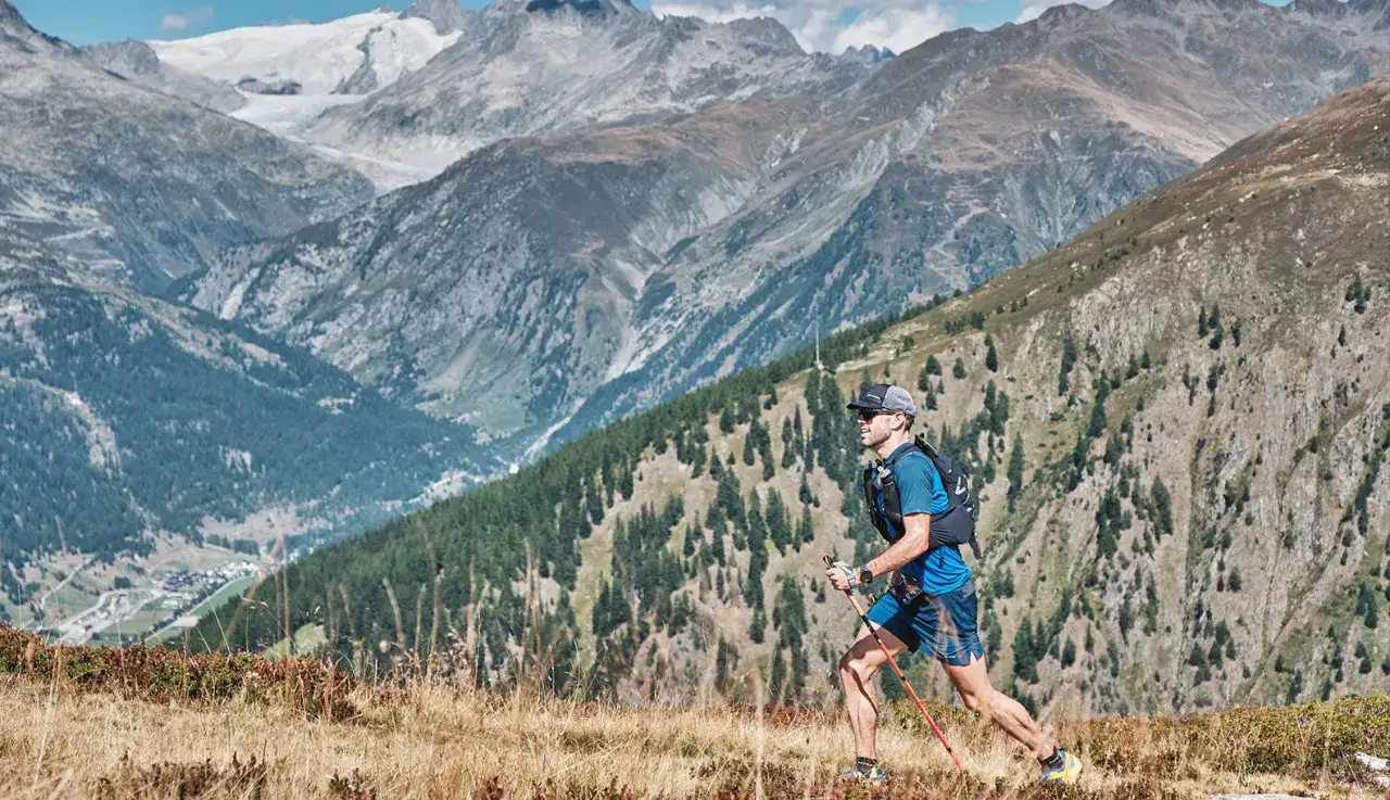 Un coureur en plein entraînement en altitude, bâtons à la main, traverse un sentier de montagne escarpé avec des sommets enneigés en arrière-plan.