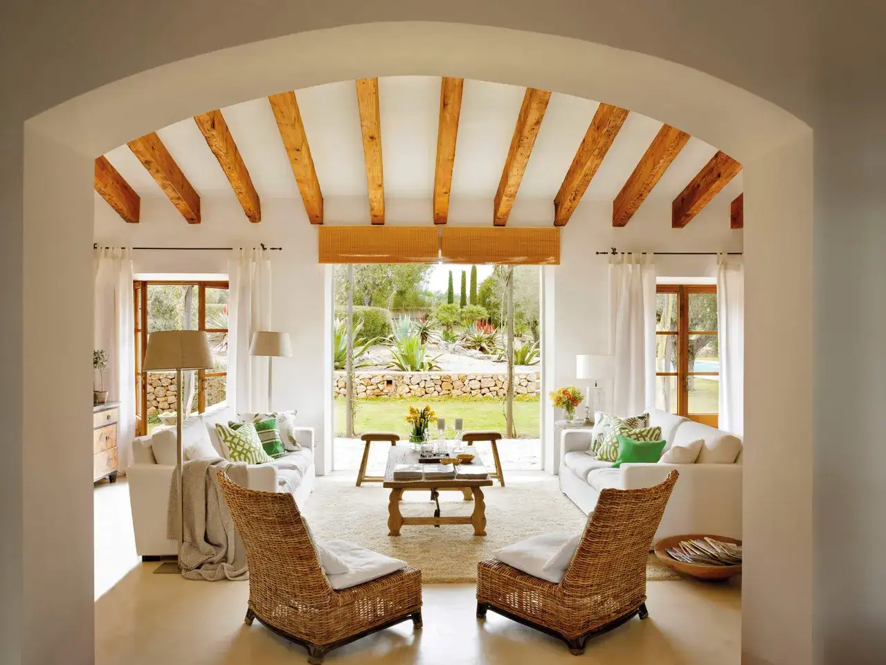 Salón con decoración ibicenca, vigas de madera, sofás blancos y sillones de mimbre, con vistas a un jardín mediterráneo.