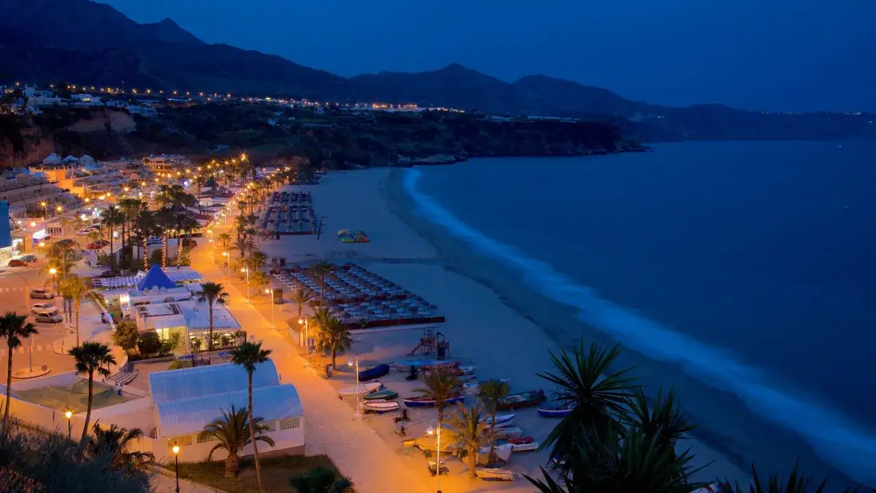 Playa de Burriana im Abendlicht: beleuchtete Promenade, Palmen, Liegen und Boote am Strand, im Hintergrund die Berge.
