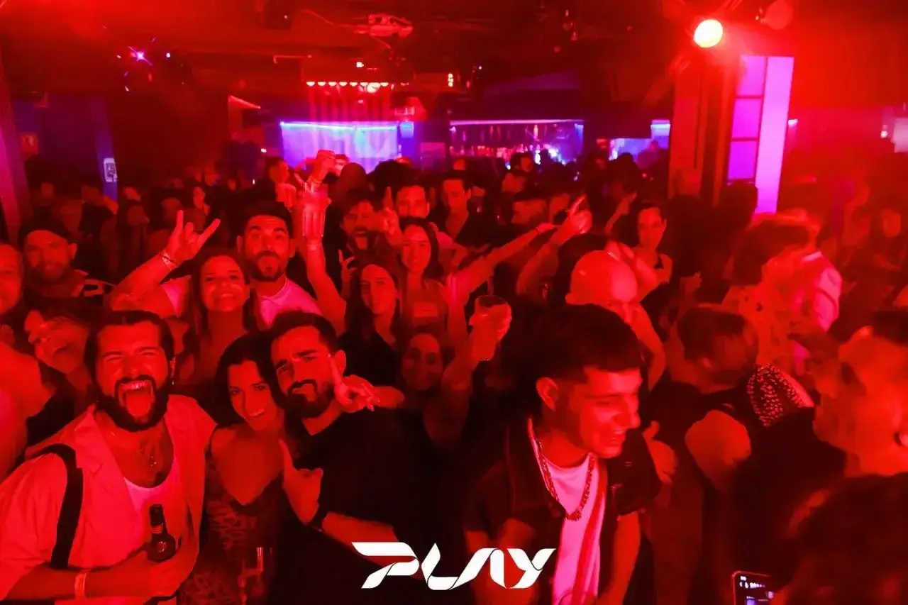 Folla festante in un locale notturno a Valencia, con luci rosse e musica. Un gruppo di amici si diverte, alcuni con drink in mano.