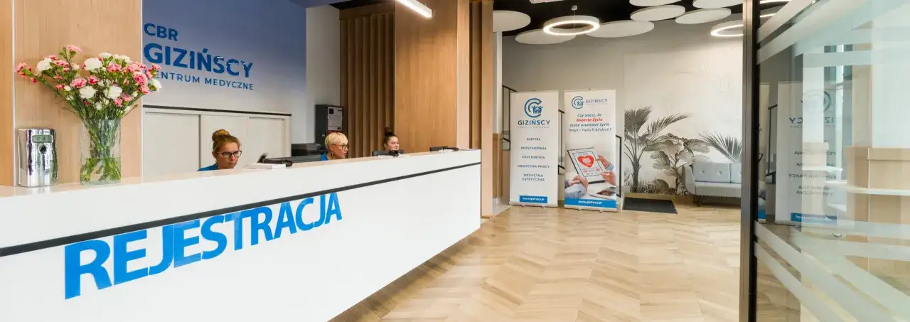 Recepcja nowoczesnego centrum medycznego z personelem, bukietem kwiatów i banerami informacyjnymi.