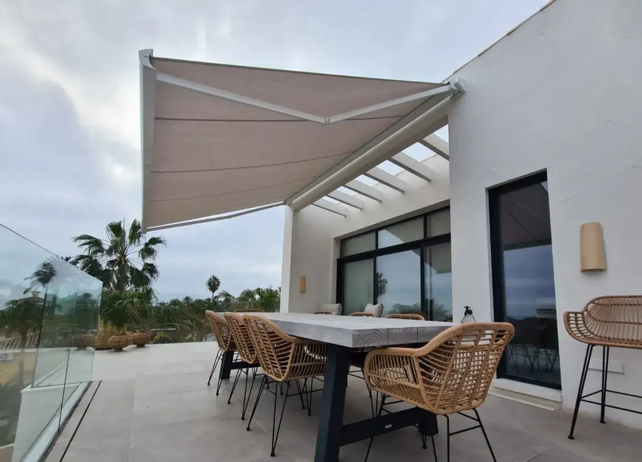 Terraza con mesa y sillas de mimbre bajo toldos extensibles. Varios tipos de toldos protegen este espacio exterior, ideal para disfrutar del paisaje.