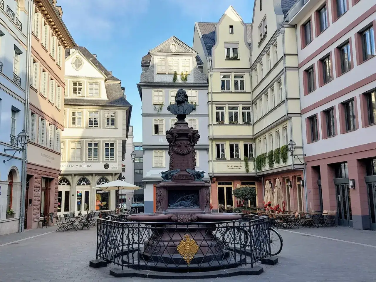 Brunnen mit Büste im Zentrum eines Platzes in Frankfurt. Bunte Häuserfassaden und Straßencafés säumen den Markt.