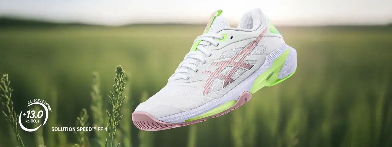 Białe buty tenisowe z różowymi i zielonymi akcentami od firmy Asics, model Solution Speed FF 4, unoszą się na tle zielonego pola.