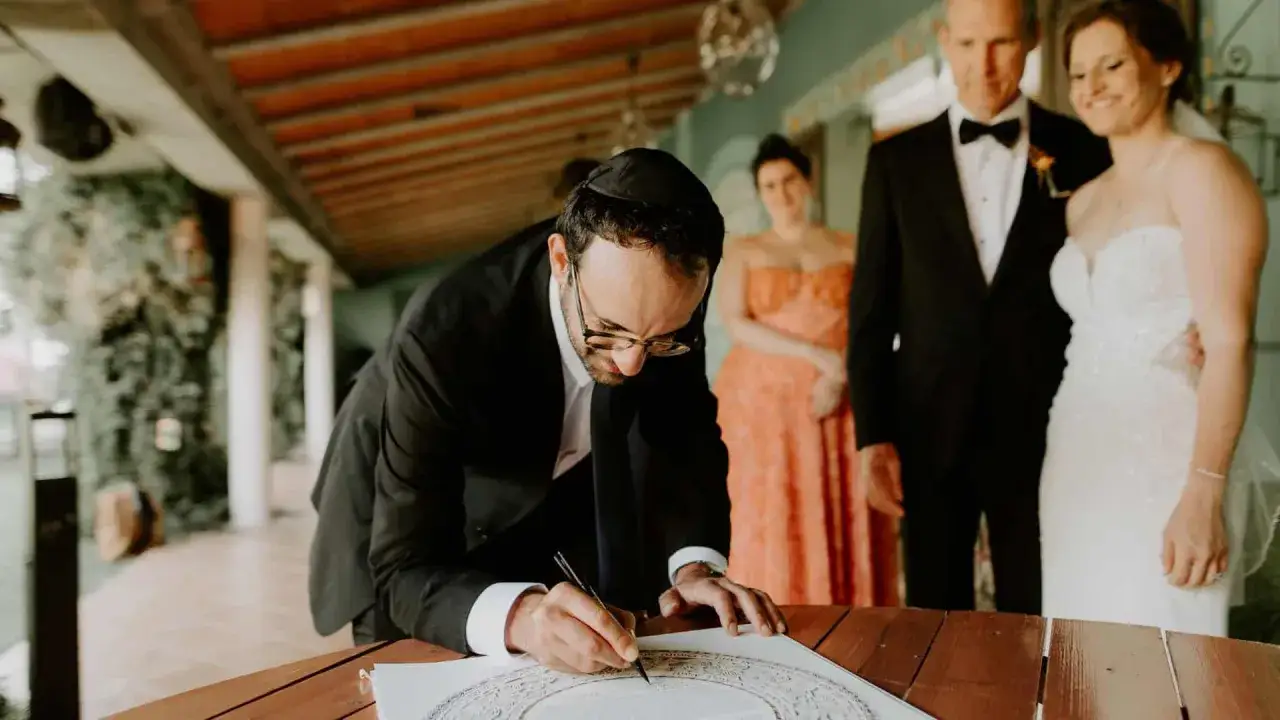 Hombre con kipá firma ketubá en un hermoso matrimonio judío. La pareja sonríe al fondo.
