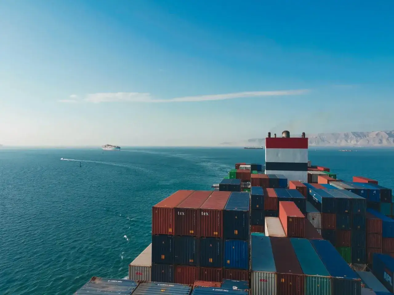 Ein riesiges Containerschiff, die CMA CGM Barracuda, fährt auf dem Meer. Berge im Hintergrund, ein kleineres Schiff im Vordergrund.