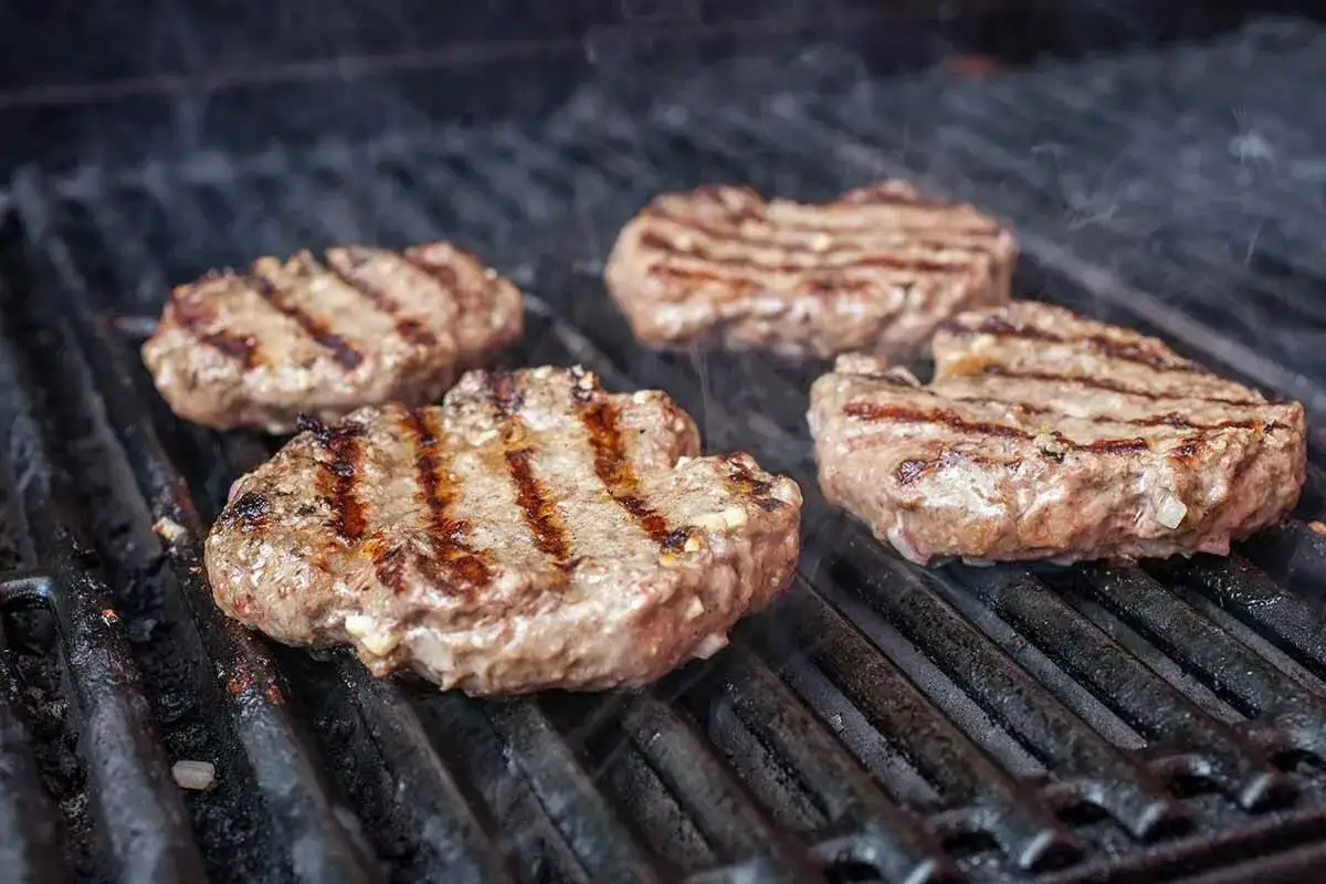 Cuatro jugosas hamburguesas caseras a la parrilla, humeantes y con marcas doradas. &iexcl;Perfectas para aprender como hacer hamburguesas caseras!