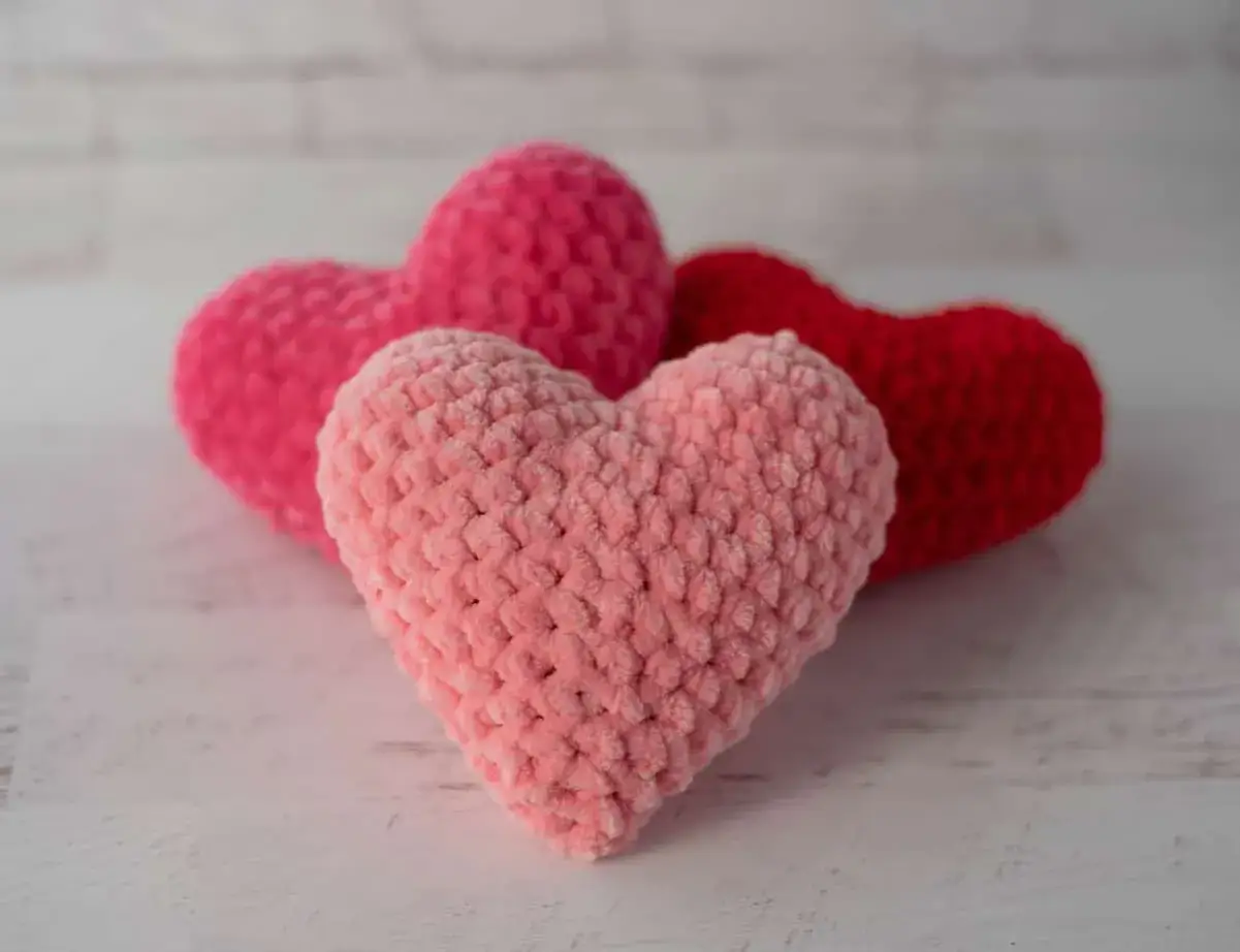 Tres corazones amigurumi de peluche, uno rosa claro al frente, otro rosa fuerte y uno rojo detr&aacute;s, sobre una superficie blanca.