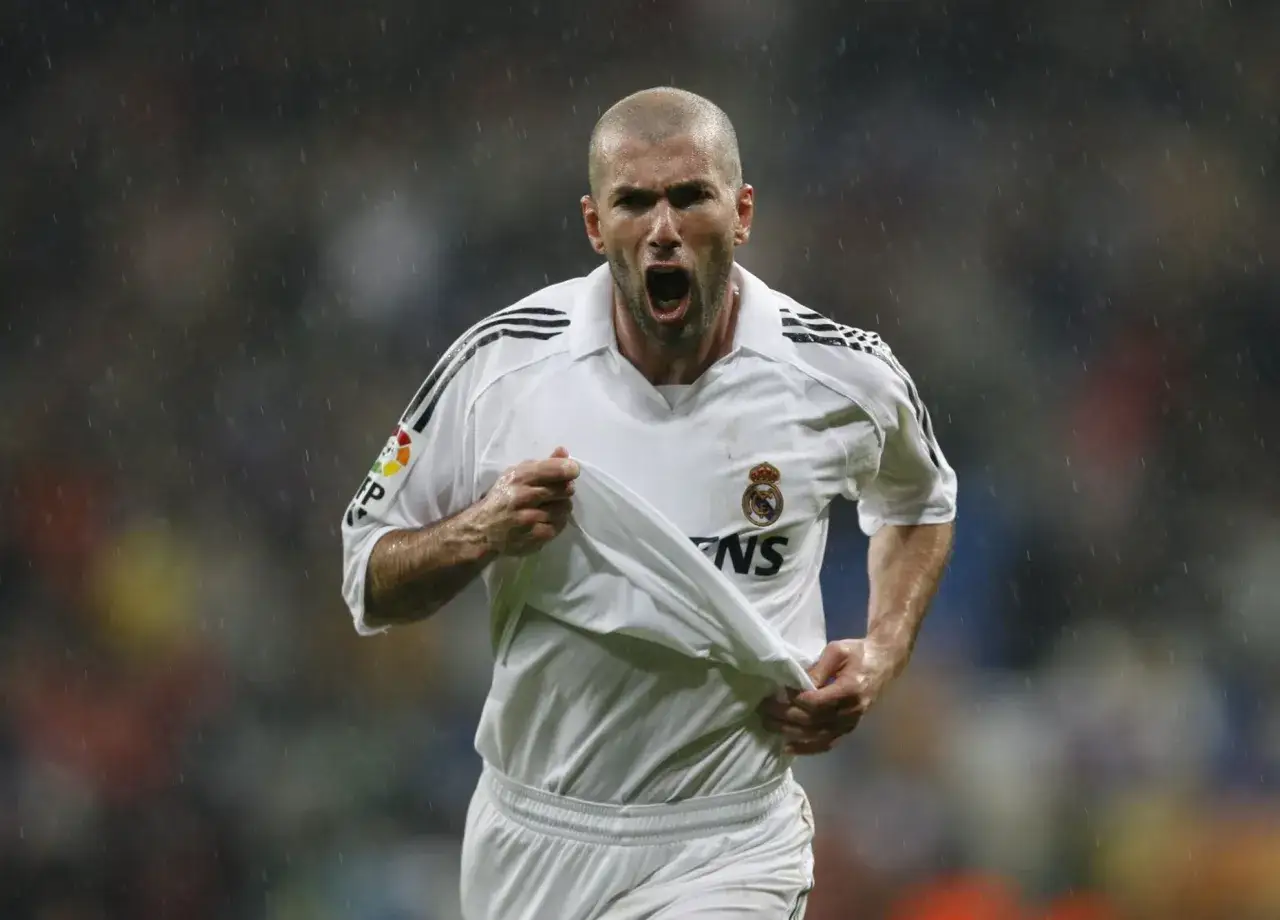 Zinedine Zidane w białej koszulce Realu Madryt, z podniesioną koszulką, krzyczy radośnie dla kibiców.