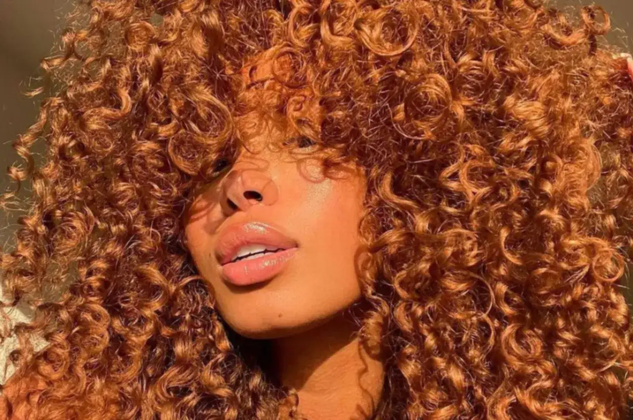 Melena voluminosa con un corte de pelo rizado a capas en tonos cobrizos. Los rizos definidos y el brillo natural resaltan la textura y el movimiento del cabello.
