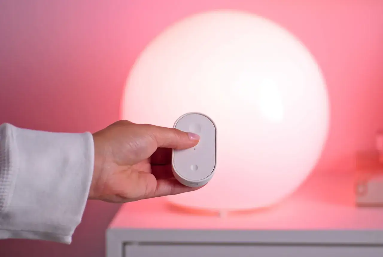 A hand holds a white remote, controlling new IoT devices. A large, glowing orb lamp casts a soft pink light.