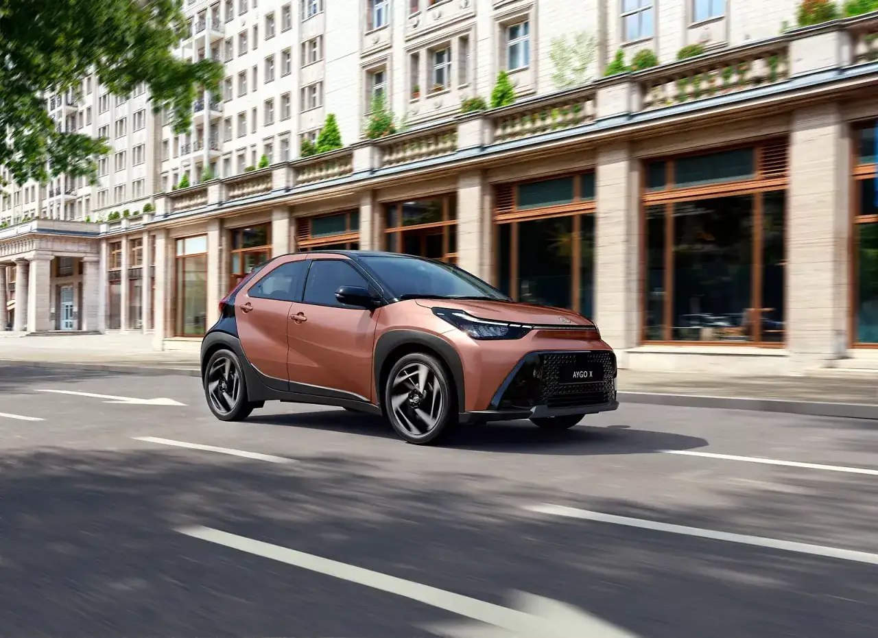 Miedziany Toyota Aygo X Hybrid sunie ulicą przed elegancką kamienicą.