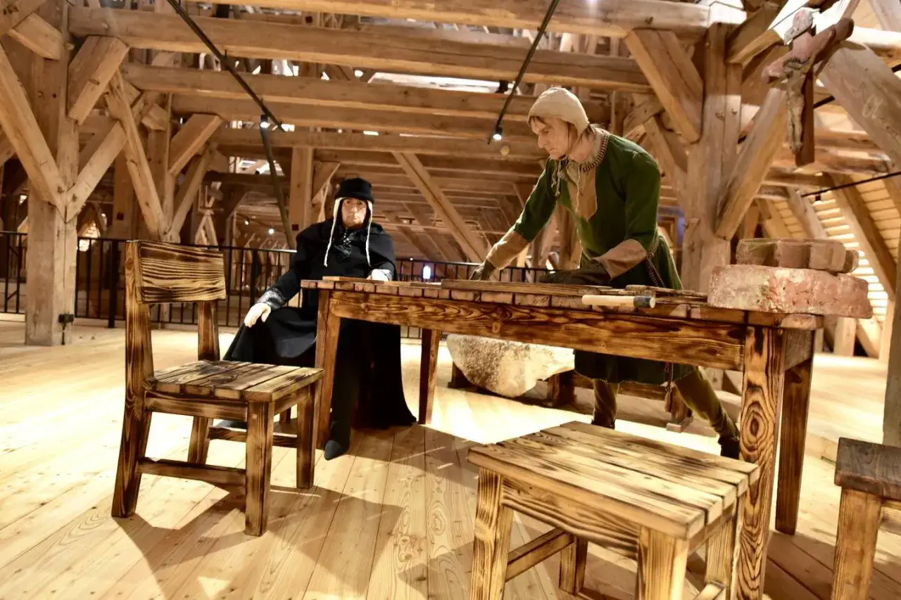 W muzeum Mazury dwie postacie w strojach historycznych przy drewnianym stole. Jedna siedzi, druga stoi, pracując.