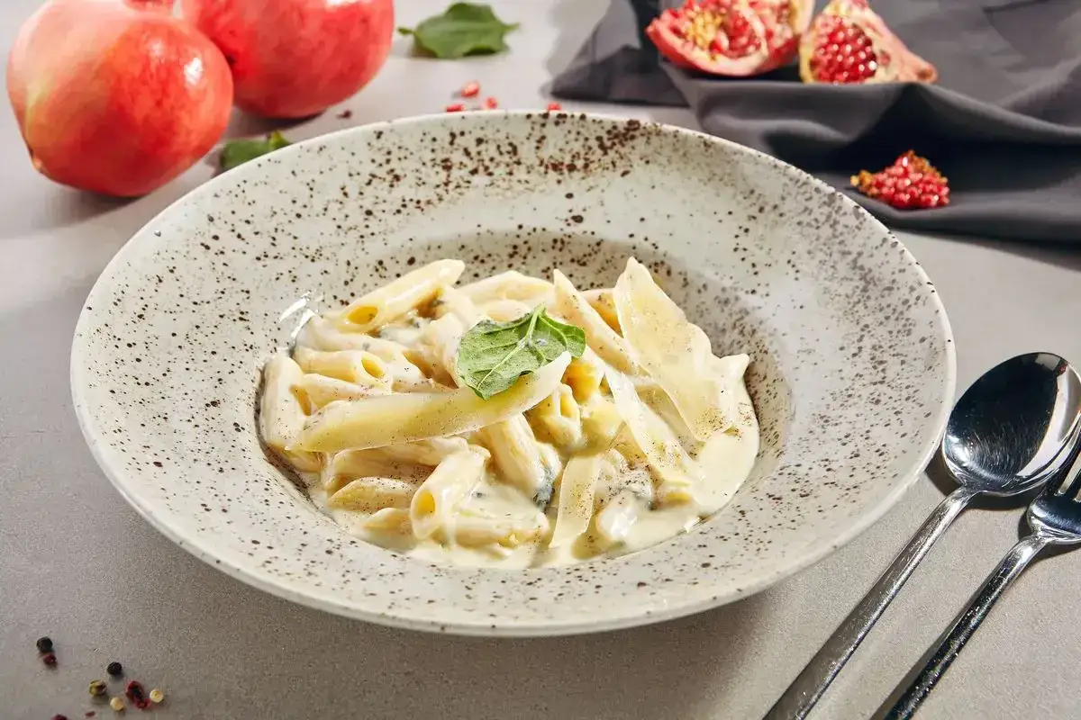 Penne cremose con una deliziosa fonduta per pasta, guarnite con scaglie di parmigiano e una foglia di basilico. Sullo sfondo, melograni freschi.