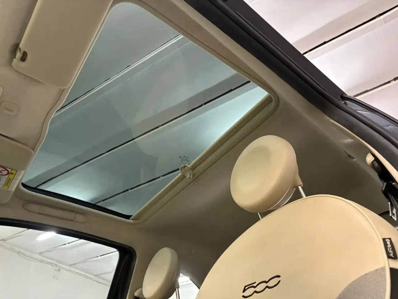 Interior de un Fiat 500 con techo solar panorámico abierto. Se ven los asientos de cuero beige y el parasol.