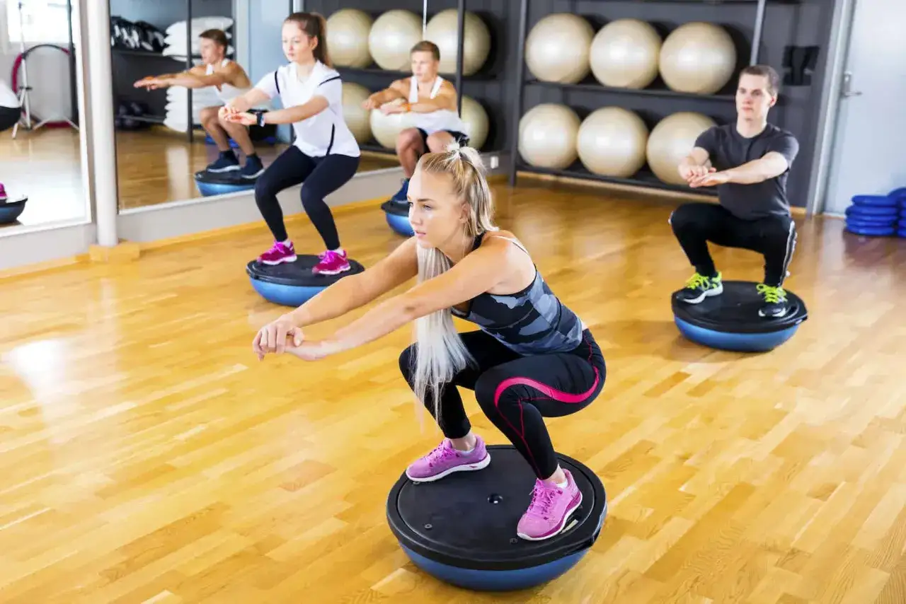 Ludzie wykonują przysiady na platformach BOSU. To świetne ćwiczenia na refluks, wzmacniające mięśnie brzucha i poprawiające r&oacute;wnowagę.