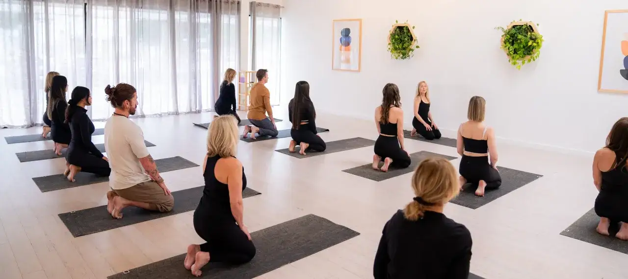 En grupp m&auml;nniskor sitter p&aring; yogamattor i en ljus studio. Vissa fokuserar p&aring; andning, andra p&aring; r&ouml;relse, en p&aring;minnelse om att pilates och yoga kan ge liknande lugn.