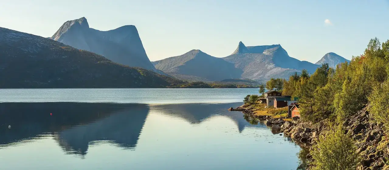 Majestätiska berg speglas i lugnt vatten vid en idyllisk kust med röda hus. Perfekt för vandring i Narvik.