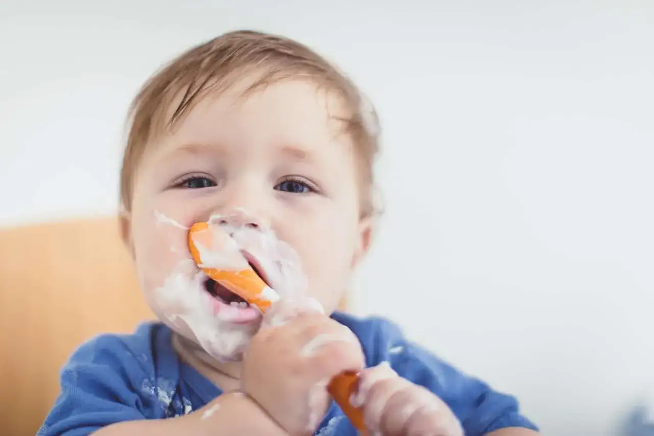 Bebé con cara y cuchara cubiertas de yogur, disfrutando de su comida. ¡Momento perfecto cuando puedo dar yogur a mi bebé!