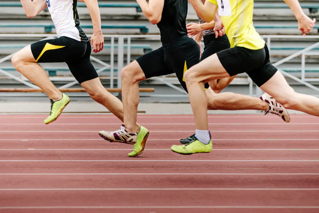 Trois athlètes en pleine course sur une piste d'athlétisme. Leurs jambes musclées et leurs pointes de course témoignent d'un renfo course à pied intense.