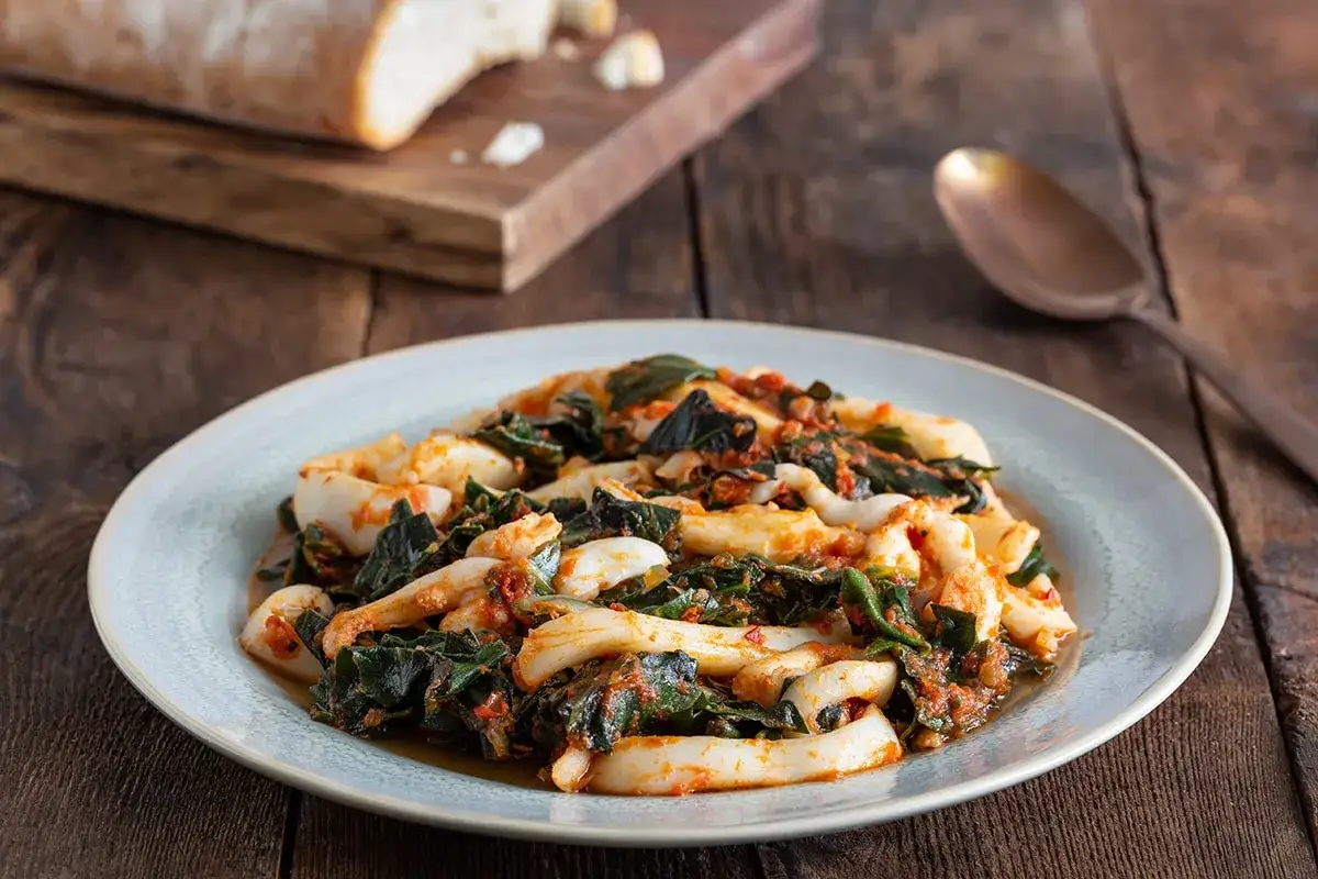 Piatto di seppie in zimino, con verdure scure e sugo rosso, su un piatto chiaro. Accanto, pane su un tagliere e un cucchiaio.