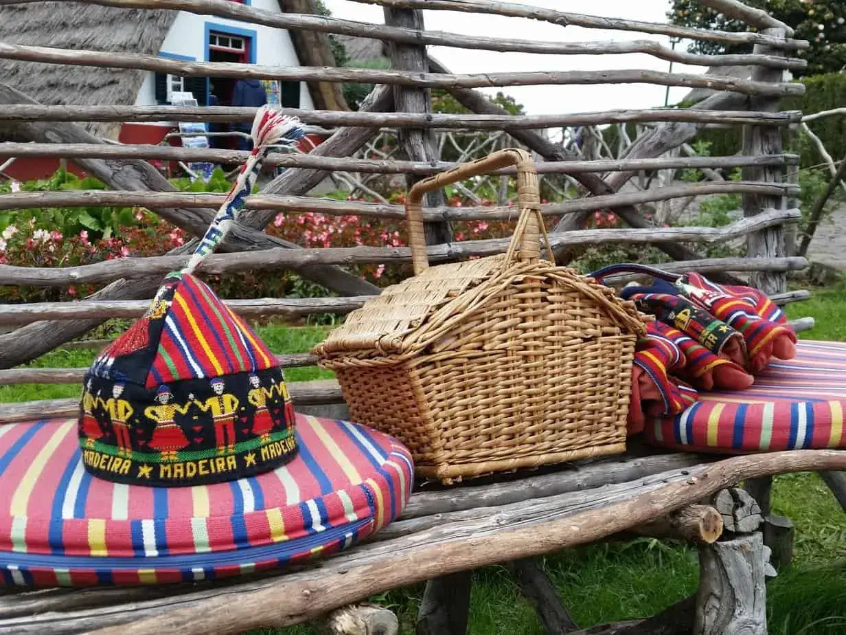 Typisch Madeira Mitbringsel: Ein bunter Hut mit Stickerei, Picknickkorb und gestreifte Kissen auf einer Holzbank vor einem traditionellen Haus.