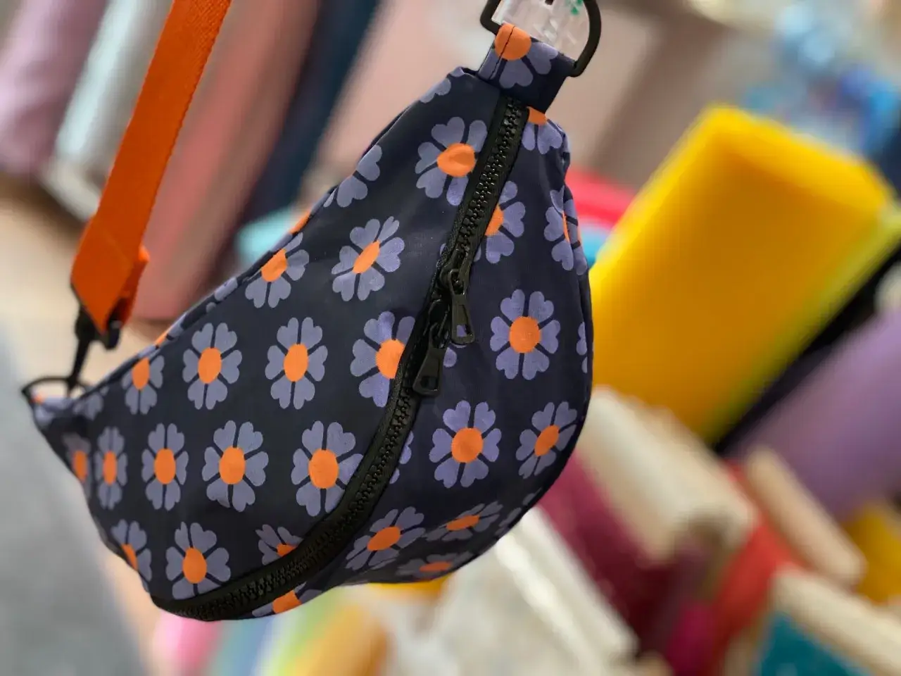 Blumige Bauchtasche mit orangefarbenem Gurt. Das Schnittmuster zeigt lila Bl&uuml;ten mit orangefarbenen Zentren auf dunkelblauem Grund.