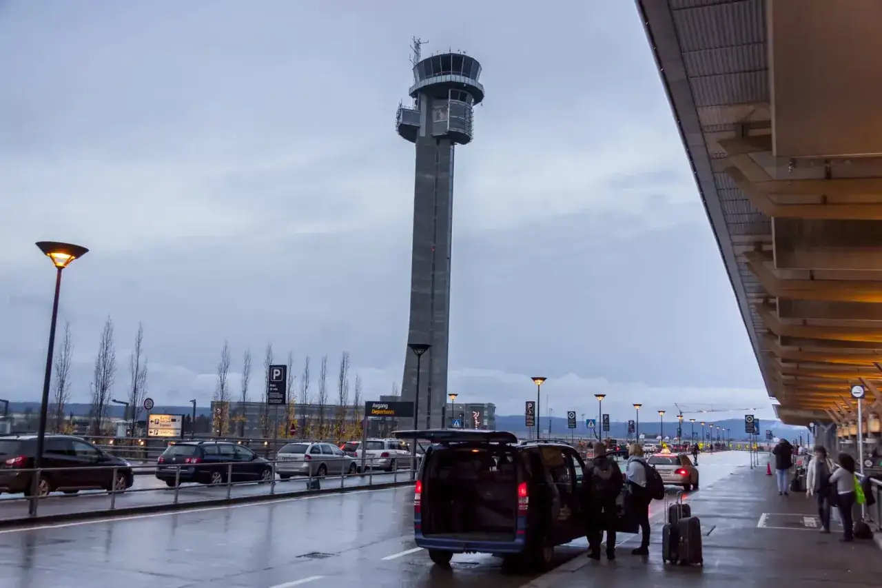 Wieczór na oslo lotnisko. Wieża kontroli lotów góruje nad mokrym asfaltem, gdzie ludzie wysiadają z vana z bagażami.