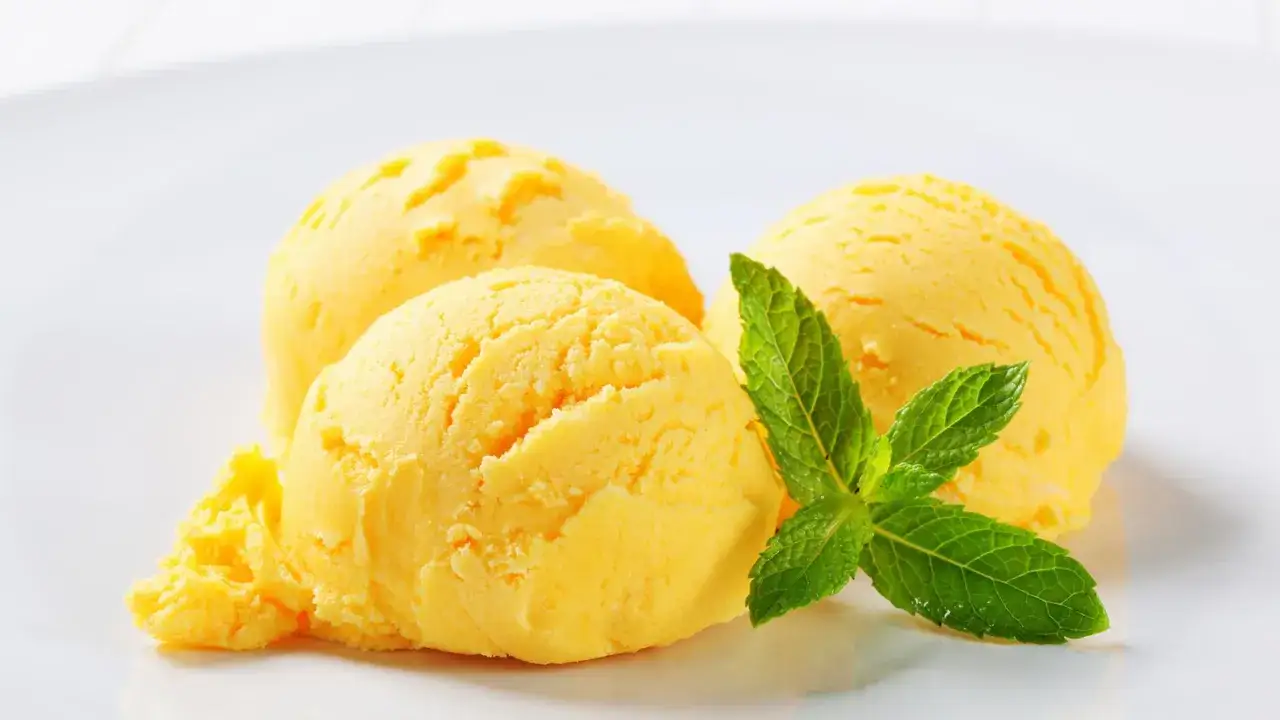 Drei Kugeln leuchtend gelbes Eis, perfekt zum orangeneis selber machen, garniert mit frischer Minze auf einem weißen Teller.