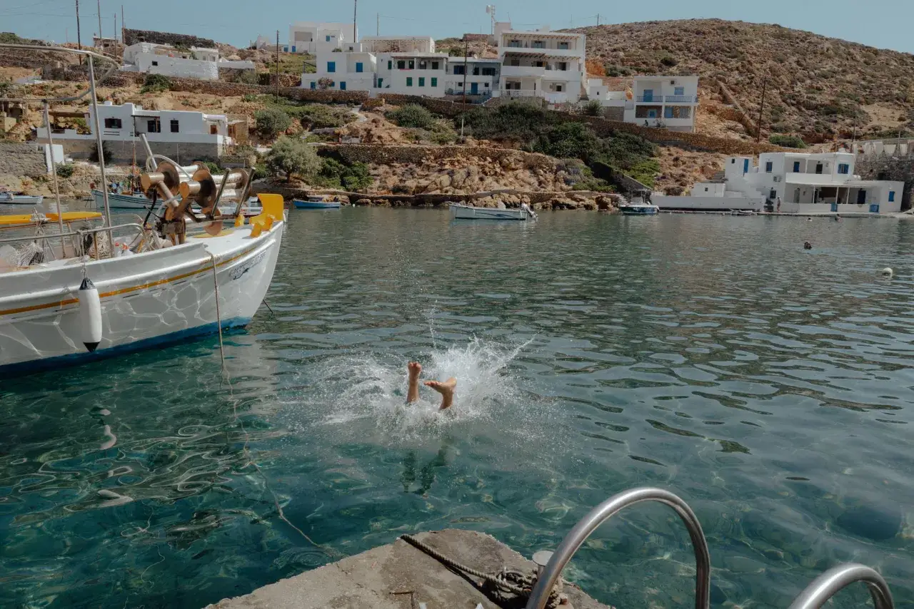 Ett barn hoppar i vattnet vid en hamn på Sifnos, Grekland. Vita hus klättrar uppför en kulle bakom en båt.