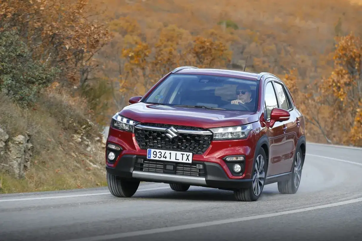 Röd Suzuki S-Cross kör på en kurvig väg i höstlandskap. En man med solglasögon kör bilen under ett suzuki s-cross test.